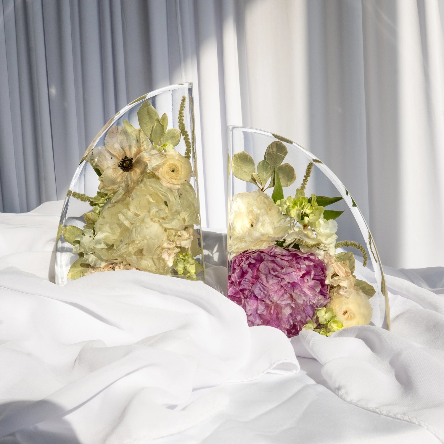Resin bookends featuring preserved wedding flowers in soft ivory, pale yellow, and plum tones, elegantly displayed on white fabric with sunlight casting soft shadows.