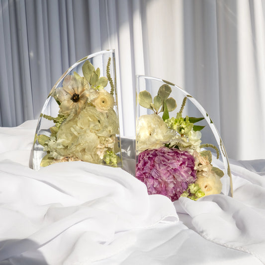 Resin bookends featuring preserved wedding flowers in soft ivory, pale yellow, and plum tones, elegantly displayed on white fabric with sunlight casting soft shadows.