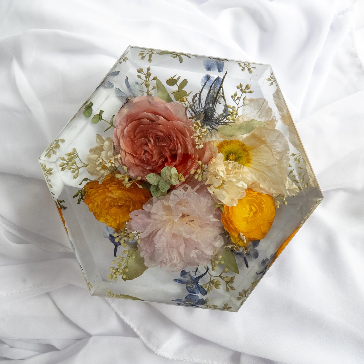 Hexagon-shaped resin floral block featuring preserved roses, ranunculus, peonies, and wildflowers in pink, yellow, and white tones on a white fabric background.