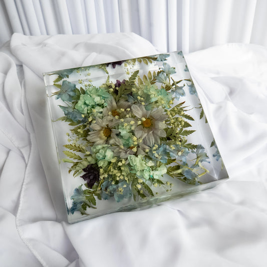 Preserved wedding flowers in a square resin block featuring white daisies, blue hydrangeas, and greenery, styled on white fabric background.