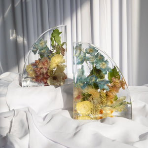 Set of resin bookends with preserved flowers in shades of blue, yellow, and peach, arranged organically and captured in warm natural light on white fabric.