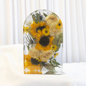Arch-shaped resin keepsake with preserved sunflowers, ivory roses, blue thistle, and yellow daisies from a wedding bouquet
