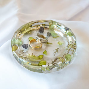 Crystal-clear resin jewelry dish with preserved green botanicals and eucalyptus embedded throughout, holding a diamond engagement ring set and gold wedding band, displayed on white fabric
