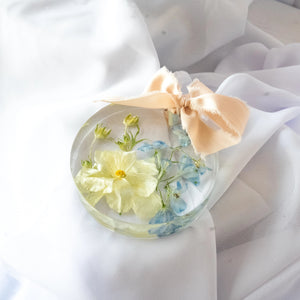Round resin ornament featuring a preserved yellow flower and soft blue blooms, finished with a blush silk ribbon — handcrafted floral keepsake by Everlasting Bloom Co.
