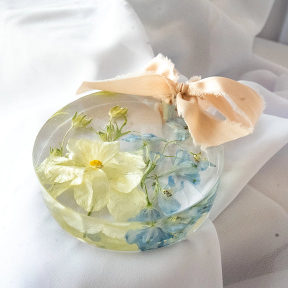 Handmade resin ornament with preserved yellow and blue wedding flowers and a blush silk ribbon, styled on white fabric — floral preservation by Everlasting Bloom Co.