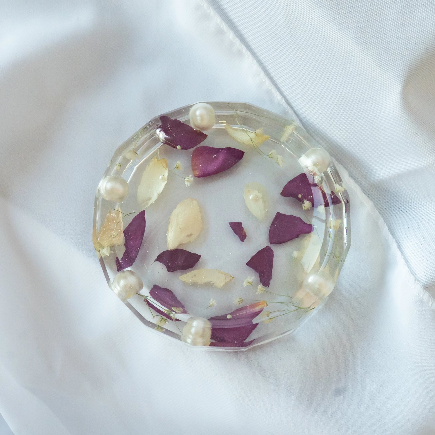 Round resin jewelry dish with faceted edges, featuring preserved purple and cream flower petals, tiny blooms, and pearl accents — wedding keepsake by Everlasting Bloom Co.