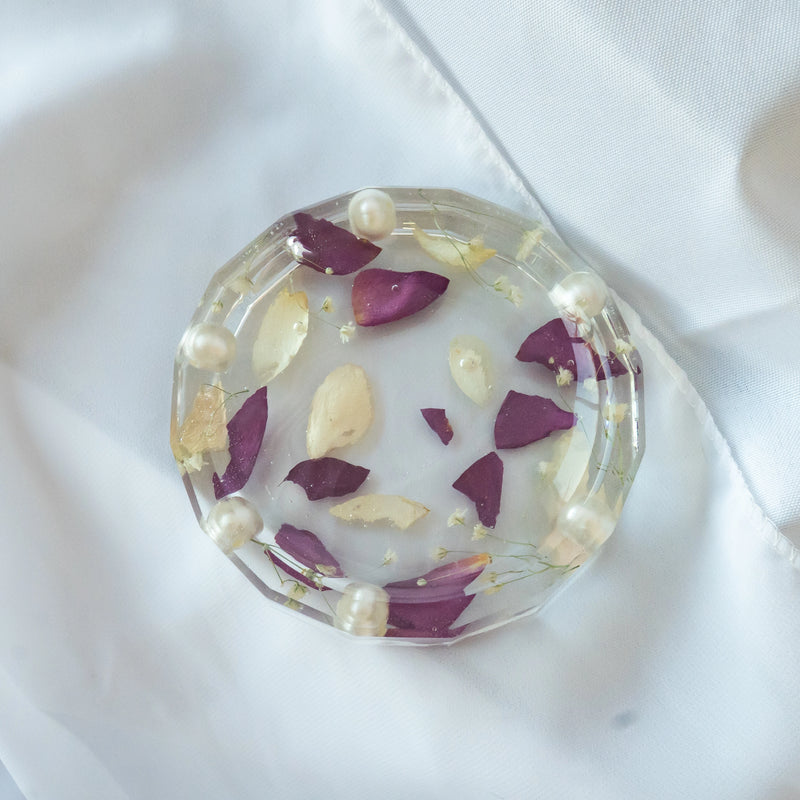 Round resin jewelry dish with faceted edges, featuring preserved purple and cream flower petals, tiny blooms, and pearl accents — wedding keepsake by Everlasting Bloom Co.