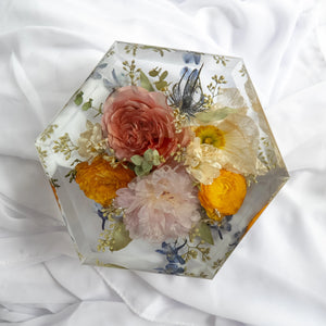 Hexagon-shaped resin floral block featuring preserved roses, ranunculus, peonies, and wildflowers in pink, yellow, and white tones on a white fabric background.