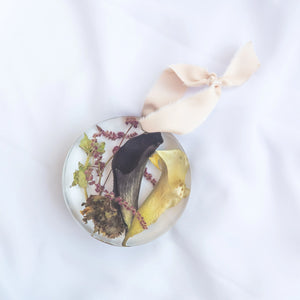 Round resin ornament with blush silk ribbon, preserved black and yellow calla lilies, burgundy astilbe, and dried thistle from a wedding bouquet