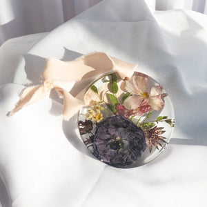 Round resin ornament with peach silk ribbon, preserved eggplant anemone, blush anemone, cream chamomile, and burgundy scabiosa from a wedding bouquet