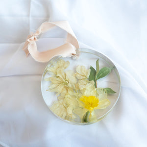 Round resin ornament with peach silk ribbon, preserved yellow daffodil, cream stock, and green foliage from a wedding bouquet