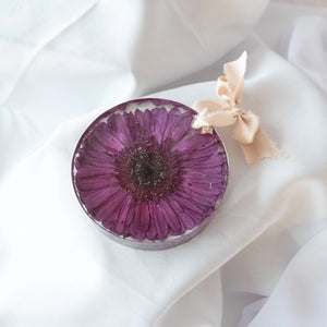 Handmade round resin ornament with a preserved purple gerbera daisy, finished with a cream silk ribbon bow, displayed on white fabric