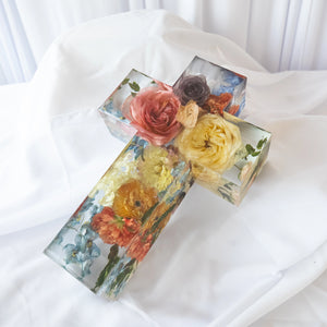Clear resin cross with preserved pink, yellow, and orange roses, blue delphinium, and greenery, shown from above on white fabric