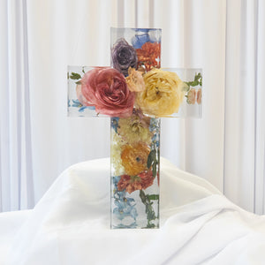 Clear resin cross with preserved pink, yellow, and orange roses, blue delphinium, and greenery, displayed upright on white fabric