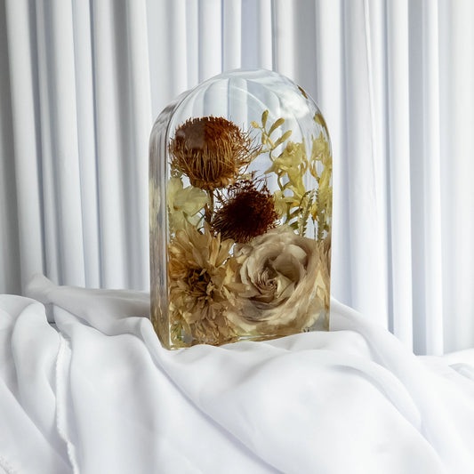 Small resin arch keepsake with preserved neutral-toned flowers, including cream roses, dried protea, and golden foliage, displayed on white fabric with a soft curtain backdrop.