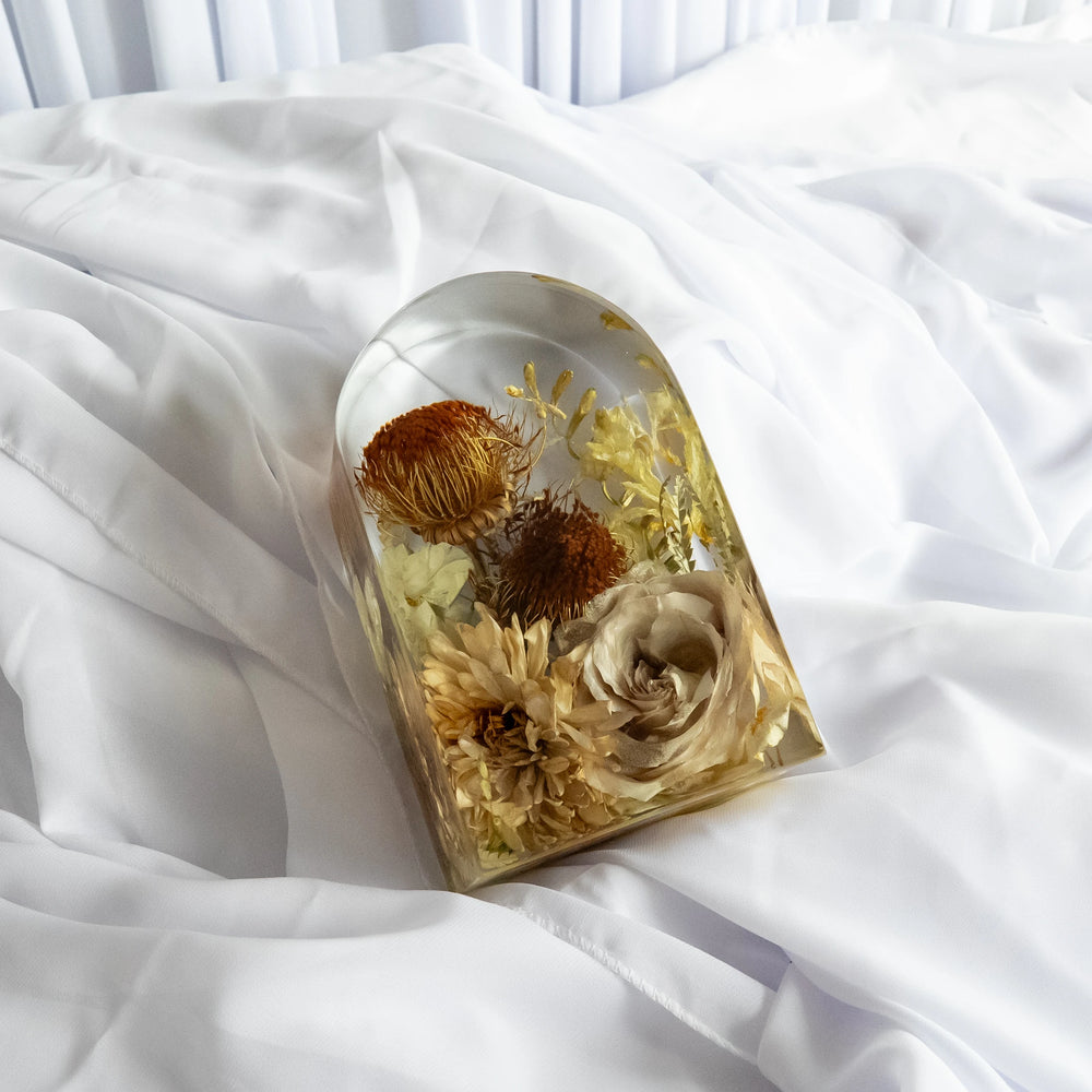 Small resin arch keepsake featuring preserved neutral-toned flowers, including roses, chrysanthemums, and protea, styled on soft white fabric.