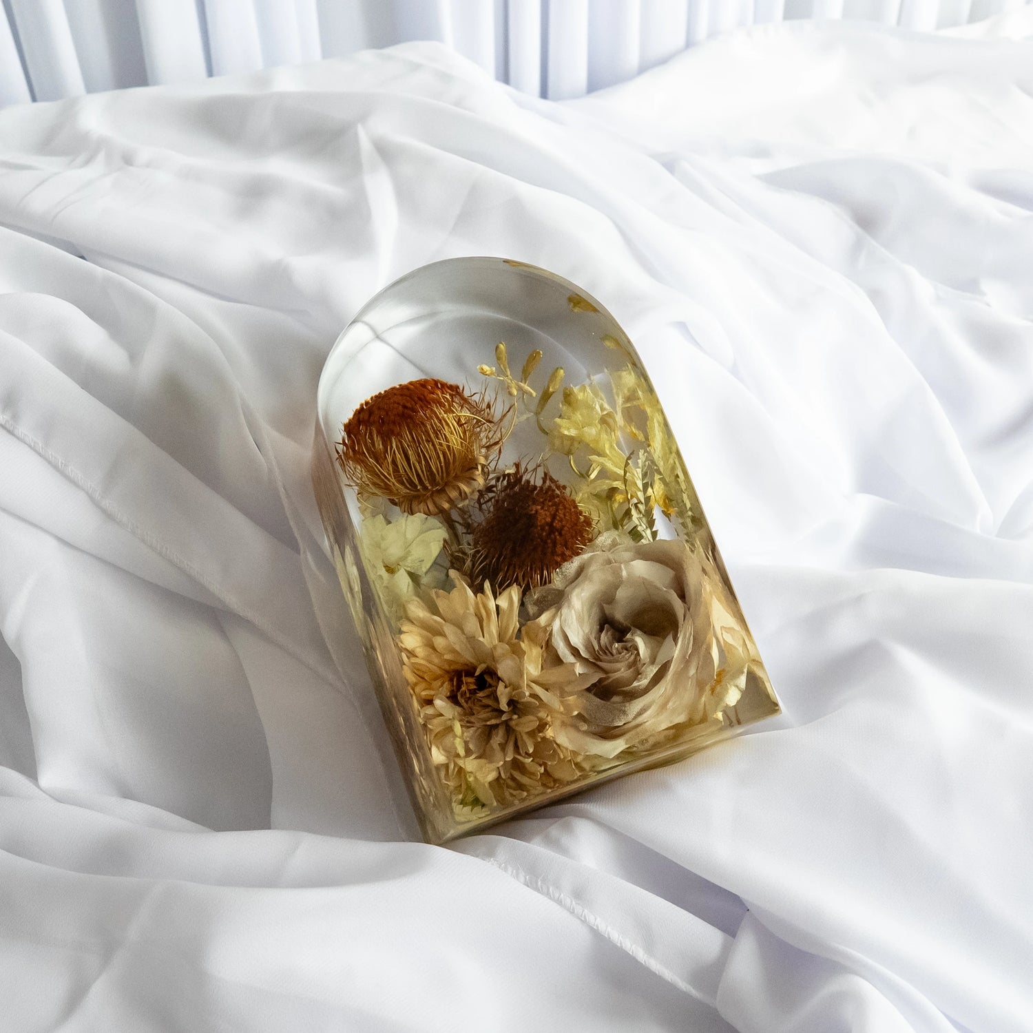 Small resin arch keepsake featuring preserved neutral-toned flowers, including roses, chrysanthemums, and protea, styled on soft white fabric.