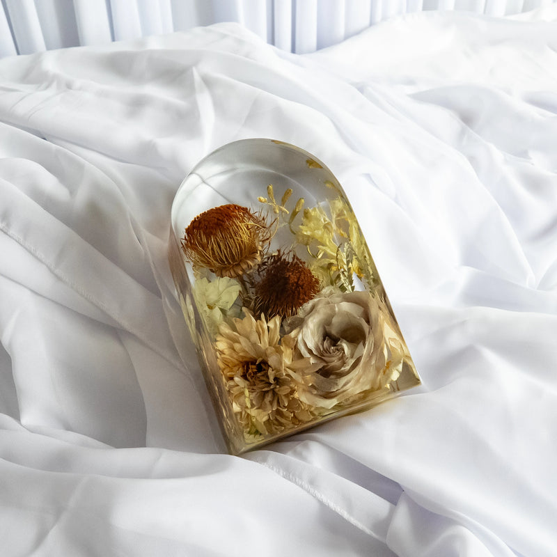 Small resin arch keepsake featuring preserved neutral-toned flowers, including roses, chrysanthemums, and protea, styled on soft white fabric.