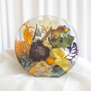 Standing circular resin display with preserved orange pincushion protea, yellow calla lily, deep purple strawflower, cream roses, and yellow craspedia from a wedding bouquet